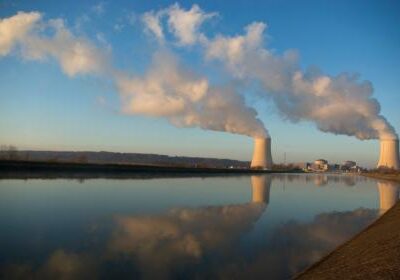 Water-reflected picture of two nuclear power plant cooling towers with steam emitting across a blue sky